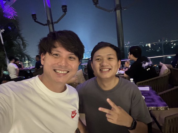 Two young men smile at the camera in a selfie taken at an outdoor rooftop venue at night, with city lights visible in the background; one wears a white Nike shirt and the other makes a peace sign.