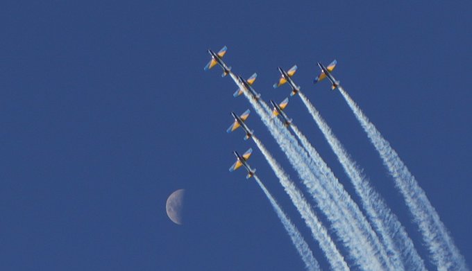 A formation of jets flying in diagonal lines with white contrails against a deep blue sky, with a crescent moon visible in the lower left.