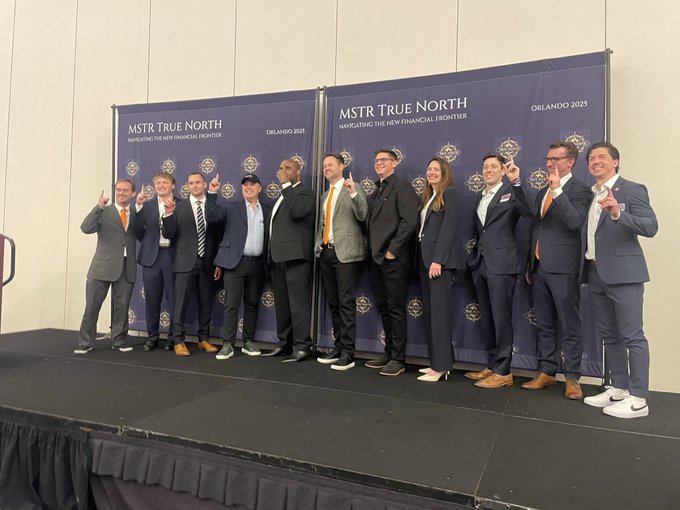 A group of professionally dressed individuals pose together on stage, flashing "₿" hand signs in front of two blue banners reading "MSTR TRUE NORTH — NAVIGATING THE NEW FINANCIAL FRONTIER — ORLANDO 2025," visually echoing the "₿uild on ₿itcoin" message and symbolizing MicroStrategy's Bitcoin-focused vision for the future of finance.