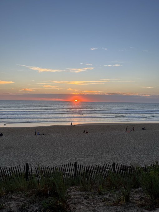 A serene sunset scene at a beach with the orange sun setting over the ocean, silhouetted figures on the sand, and a wooden fence with dune grass in the foreground.
