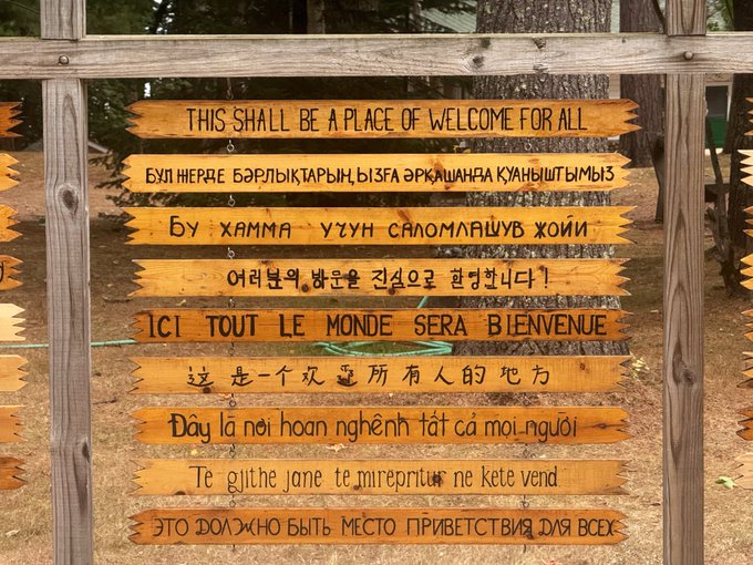 A rustic wooden sign displays a welcome message in multiple languages, prominently reading "THIS SHALL BE A PLACE OF WELCOME FOR ALL" in English at the top. A simple "gm" (good morning) accompanies the image, greeting viewers with the universal theme of hospitality and inclusivity.
