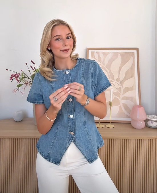 A woman wearing a denim short-sleeve top and white pants poses in a well-decorated room with floral arrangements and a framed art piece behind her.