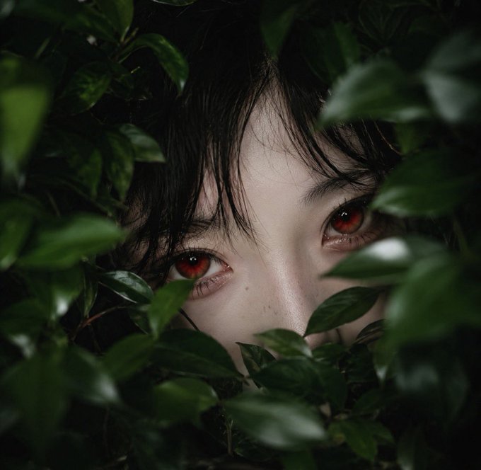A close-up portrait photograph of a person with dark hair and striking red eyes, partially obscured by green leaves in the foreground.