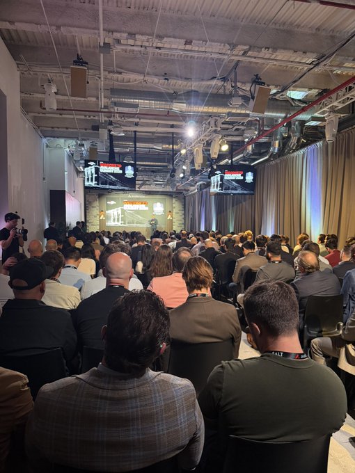 An audience sits in a dimly lit industrial-style venue watching a presentation on stage with screens displaying logos.