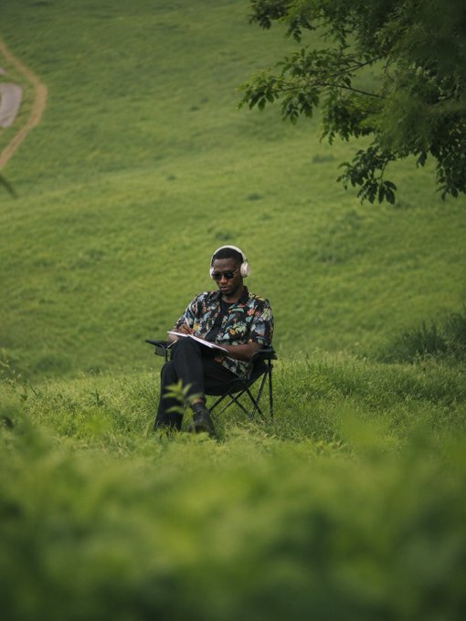 A person wearing headphones and sunglasses sits on a camping chair in a grassy field, writing or sketching on a notepad.