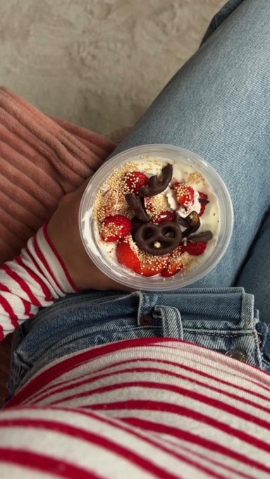 A first-person view of a person wearing a red and white striped shirt and blue jeans, holding a clear plastic cup filled with a yogurt parfait topped with strawberries, Oreos, and sprinkles.
