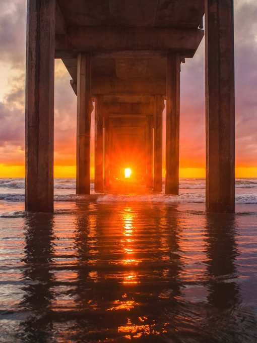 A scenic view of a pier at sunset, with the bright orange sun glowing through the structural pillars and reflecting vividly on the rippling ocean water below.