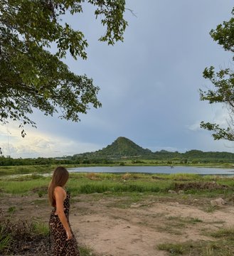 A scenic view of a woman standing on a dirt path looking out over a lake with a small mountain in the distance under a cloudy sky, framed by tree branches.