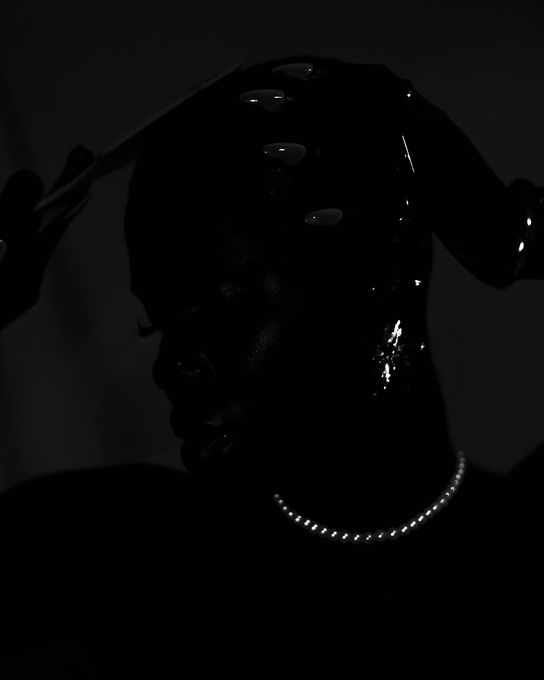 A dark, dramatic portrait photograph of a woman with wet-look hair, wearing a pearl necklace, with light catching her profile and hair accessories.