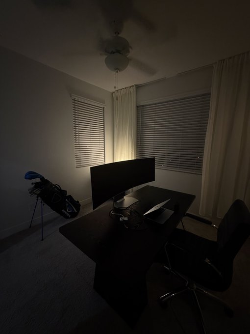 A dimly lit home office featuring a desk with a monitor, a laptop, an office chair, a ceiling fan with a light, and window blinds with sheer curtains.