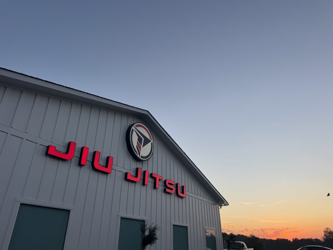 A Jiu Jitsu gym exterior with a red "JIU JITSU" sign and logo, captured against a sunset sky.
