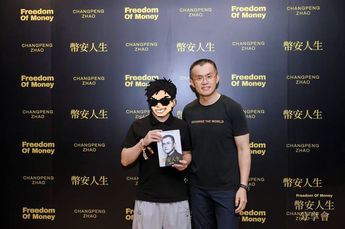 Two men posing in front of a black backdrop with 'Freedom Of Money' and 'CHANGPENG ZHAO' printed repeatedly, one holding a book featuring the other's image.