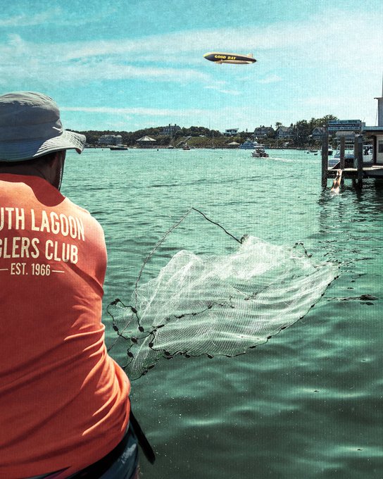 A person wearing an orange 'SOUTH LAGOON ANGLERS CLUB — EST. 1966 —' shirt throws a cast net into the water, with a Goodyear blimp flying overhead.