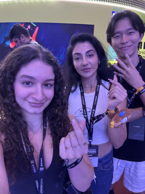 Three smiling young people pose for a selfie at what appears to be a crypto/web3 conference, wearing lanyards with "BLOCK & LOTUS" and "EVENTUS" branding along with "STUDIO" visible on the rightmost person's shirt, with a TED-style screen in the background. The image relates to the post by showing the creator enjoying herself at an industry event alongside others, presumably including someone she admires, conveying confidence and excitement rather than any negativity from critics.