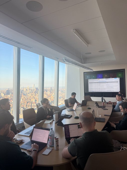 A group of professionals gather around a conference table in a high-rise meeting room with sweeping city views, working on laptops while a large screen displays a spreadsheet. The "war room" atmosphere is palpable—people sit quietly focused on their screens, likely tracking data in preparation for the "Joe's pizza mainnet" launch mentioned in the caption.