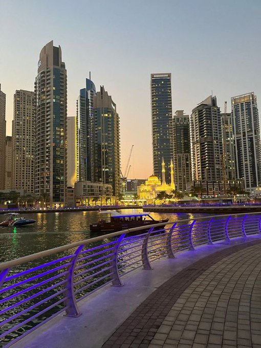 A twilight cityscape view from a waterfront promenade with purple-lit railings, showing tall skyscrapers and a glowing mosque in the distance, with boats on the water.
