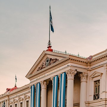 A grand neoclassical government building is adorned with blue and white striped drapery, with the Salvadoran flag flying prominently atop its columned façade. The architecture, likely an official government institution in El Salvador, visually reinforces the post's context about the country's monetary policy and Bitcoin accumulation efforts.
