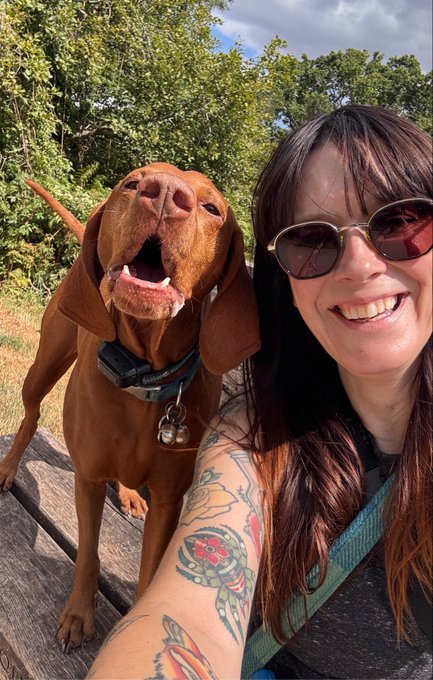 A selfie of a smiling woman with dark hair and sunglasses, showing tattooed arms, posing with a happy reddish-brown dog, likely a Vizsla, outdoors with trees in the background.