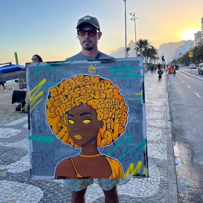 A man wearing a cap and sunglasses holds up a square canvas featuring graffiti-style artwork of a woman with an afro made of yellow text bubbles.