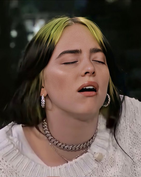 A woman with dark hair featuring yellow-green highlights sings with her eyes closed, wearing a white sweater and a diamond necklace.