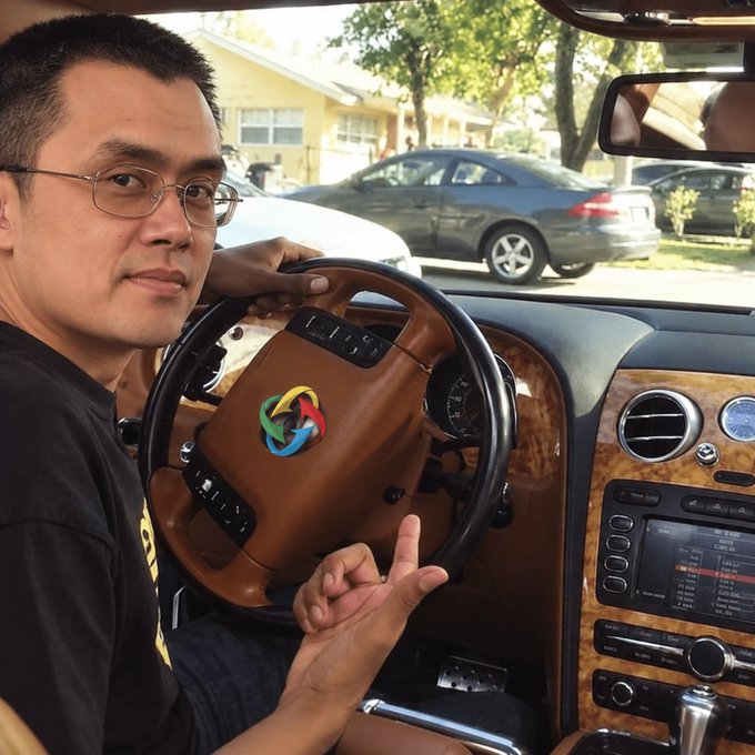 A selfie-style photo of a man with glasses sitting in the driver's seat of a luxury car with a wooden steering wheel and a colorful logo in the center.