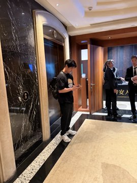 A man wearing a black t-shirt with "Aster" branding stands looking at his phone in an upscale lobby with dark marble walls, while two people in formal business attire converse in the background. The "Aster" logo and text on the man's shirt visually confirm the caption's claim of an encounter with the CEO of Aster Labs before a Binance event in Hong Kong.