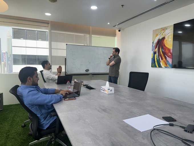 Three men gather around a white board in a modern conference room with a city skyline view, engaged in a discussion while one works on a laptop, with the "ZIGChain" reference visible in the accompanying post's mention of a collaborative brainstorming session. The room features a colorful abstract painting, a wall-mounted TV, and a meeting table with documents and electronics, suggesting an active planning meeting for the ZIGChain x Zamanat partnership.