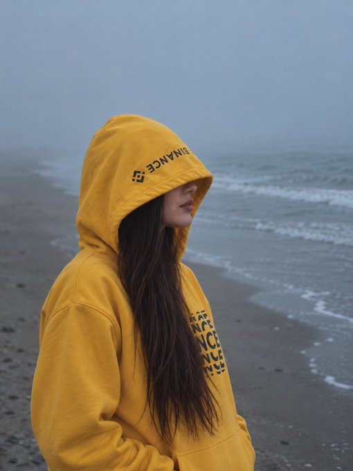 A moody portrait of a young woman wearing a yellow 'BINANCE' hoodie standing on a foggy beach with grey skies.
