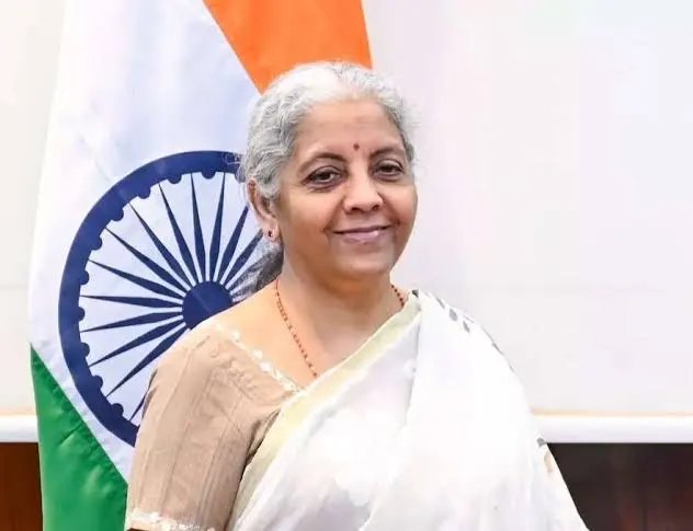 A woman with gray hair, wearing a white sari with a beige blouse and a red bindi, stands smiling in front of the Indian national flag, identifiable by its tricolor stripes and the Ashoka Chakra visible behind her. The image relates to the accompanying post, which references FM @nsitharaman's appeal to the RBI regarding CBDC pilots and digital rupee tokenisation, indicating she is likely the Finance Minister of India, Nirmala Sitharaman, pictured in an official capacity amid discussions on financial digitalization.