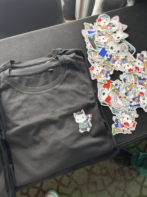 A stack of dark gray t-shirts featuring an adorable sticker of a French bulldog holding a flag sits beside a pile of colorful cartoon-style bulldog stickers, ready for the booth. The merch setup perfectly invites visitors to "come meet us at our booth!"