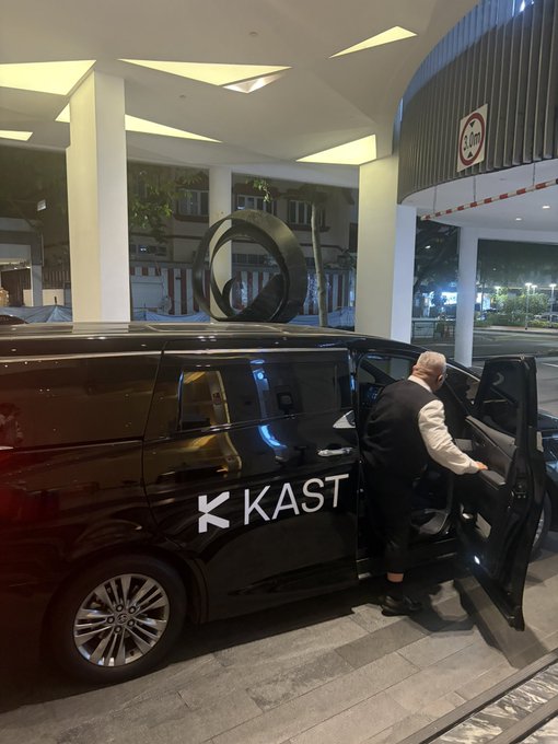 A chauffeur in a vest holds open the door of a black vehicle branded with 'KKAST' logo, parked at what appears to be a building entrance at night.