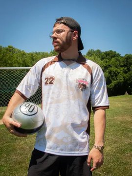 A man wearing a black cap and the white/brown 'Chimpers' football jersey (#22) holds a football on a grass field.