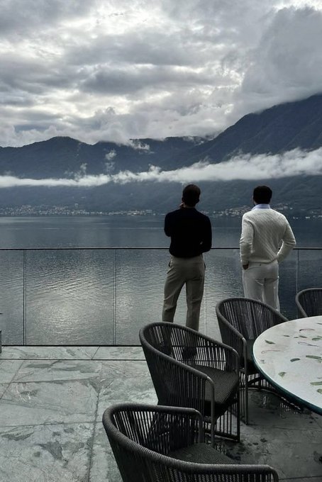 Two men standing at a railing overlooking a serene lake with mist-covered mountains in the background, taken from a patio with wicker chairs and a marble table.
