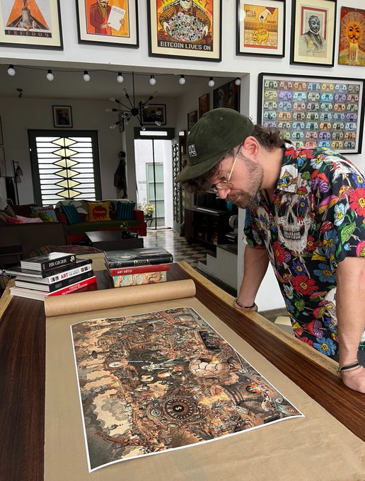 A bearded man in a colorful floral shirt and green cap looking down at a large poster or artwork being unrolled on a wooden table, in a room decorated with numerous framed Bitcoin and cryptocurrency-related posters on the walls.