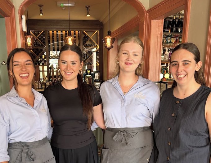 Four young female staff members, some in light blue shirts and others in black, smile and pose together behind a bar counter in a warmly lit establishment.