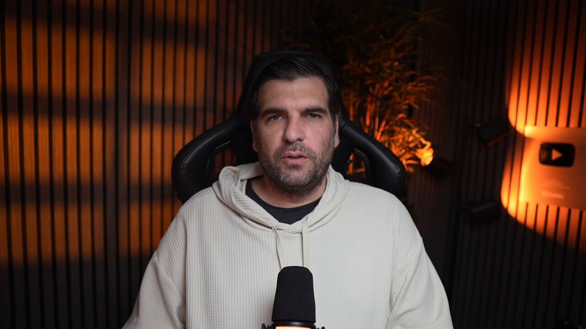The image shows a man wearing a white hoodie sitting in front of a microphone with a computer screen behind him. The background features a wooden wall with orange lights that cast an interesting glow over the scene, creating a warm atmosphere. The man appears to be speaking into the microphone or presenting something on the computer screen, possibly related to wealth or financial analysis.