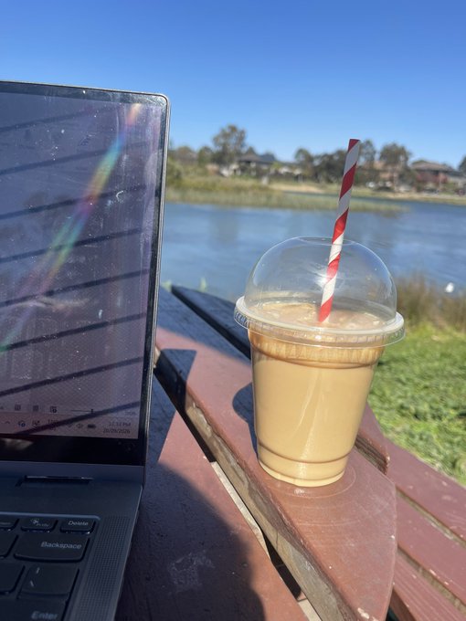 An iced coffee in a clear plastic cup with a red-and-white striped straw sits on a wooden table next to an open laptop, overlooking a lake with houses in the distance on a sunny day.