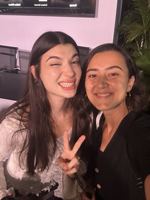 A selfie of two smiling women, one making a peace sign and a winking face, posing together indoors.