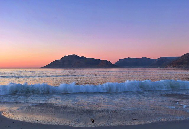 A serene beach scene at sunset with gentle waves rolling onto the shore and silhouetted mountains against a soft pink and blue sky.