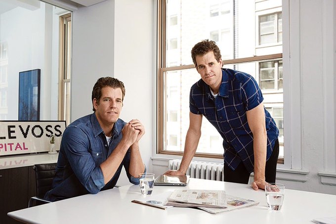 The image features two men sitting at a table, with one man wearing a blue shirt and the other wearing a plaid shirt. They are both looking directly at the camera while posing for the photo. The background of the image shows an office setting with a window behind them. There is also a sign in the background that reads "Winklevoss twins have heavily backed and supported $ZEC once again after the recent bug scare". This suggests that the Winklevoss twins are promoting Bitcoin, which could be related to the current post about privacy coins on their website.