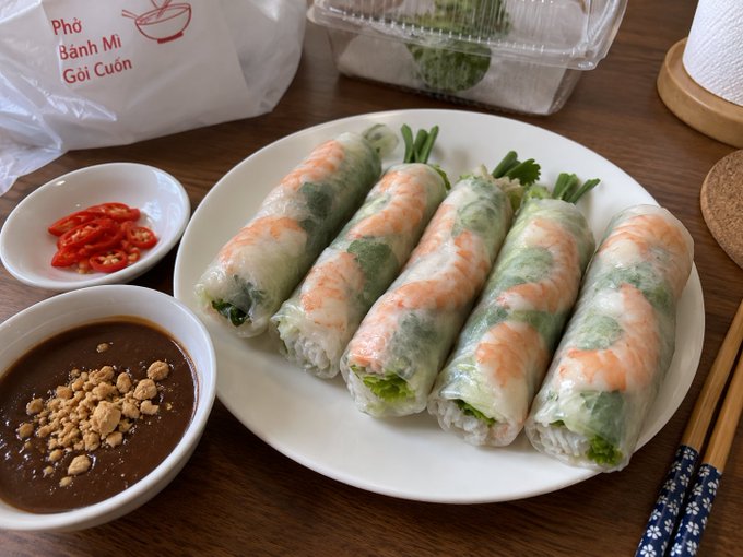 A plate of Vietnamese summer rolls (Gỏi Cuốn) filled with shrimp and greens, accompanied by a peanut sauce and sliced chili, with packaging labeled 'Phở, Bánh Mì, Gỏi Cuốn'.