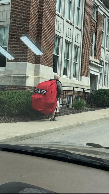 A man walking on a sidewalk in front of a brick building while carrying a large red Sansom delivery bag, photographed from inside a vehicle.