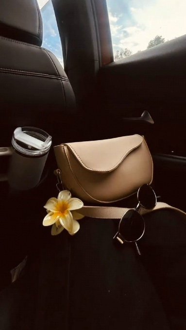 A tan leather crossbody bag sits on a dark car seat next to sunglasses, a coffee tumbler, and a yellow and white plumeria flower.