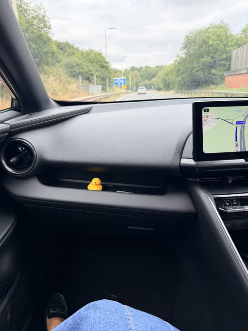 A view from the passenger seat of a car on a highway, featuring a small yellow rubber duck sitting on the dashboard near the GPS navigation screen.