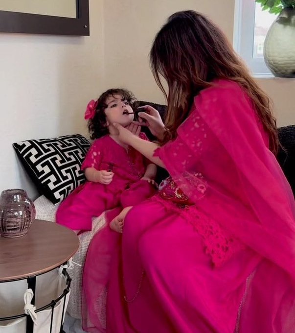 A mother in a vibrant magenta outfit carefully applies lip color to her young daughter, who is dressed in a matching pink traditional outfit and adorable matching pink bow. The tender, coordinated bonding moment between mother and child captures exactly what the poster wants in life—sweet, shared moments with her little one.