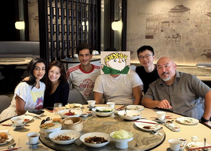 Six friends share a meal around a large round table laden with Chinese dishes, including dim sum, rice bowls, and vegetables, in what appears to be an upscale restaurant. One man in the center wears a t-shirt featuring the iconic Pudgy Penguins penguin logo, tying the gathering to the post's celebration of friendships formed through the Pudgy Penguins community.