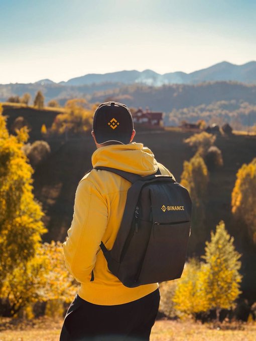 A person in a yellow hoodie and black cap wearing a black backpack with the "BINANCE" logo gazes out over a scenic mountain landscape, embodying the post's message of taking time to "touch grass" and recharge before the trading week begins.