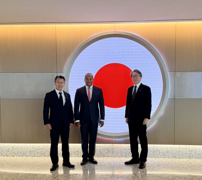 Three men in dark suits stand in front of a large illuminated Japanese flag displayed on a wall, posing for a formal photo.