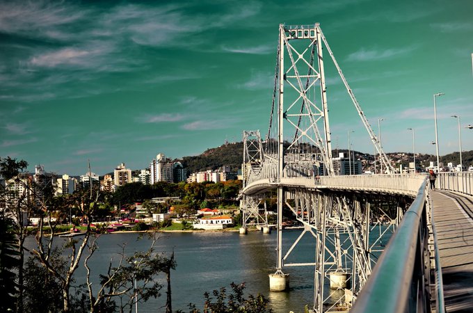 A photograph of a large steel suspension bridge spanning a body of water, with a cityscape and green hills visible in the background under a teal-tinted sky.