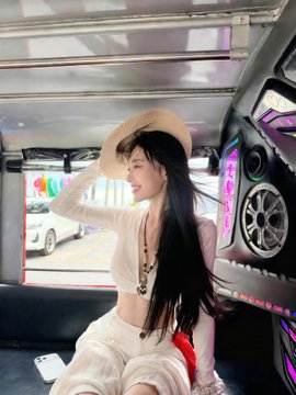 A young woman with long dark hair, wearing a beige wide-brimmed hat, a light cardigan, and a necklace, sits smiling inside a vehicle (likely a tuk-tuk or similar ride) with her hand raised near her hat. The "#binance #BinanceVip" hashtags suggest the photo is tied to a Binance VIP-related post, though no visible text or logos appear in the image itself, making the caption purely focused on the stylish portrait of the woman.